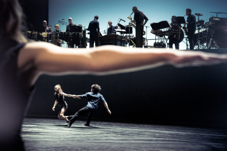 There is an orchestra at the top of the image, divided from two dancers below, by a stretched out arm framed in the foreground.