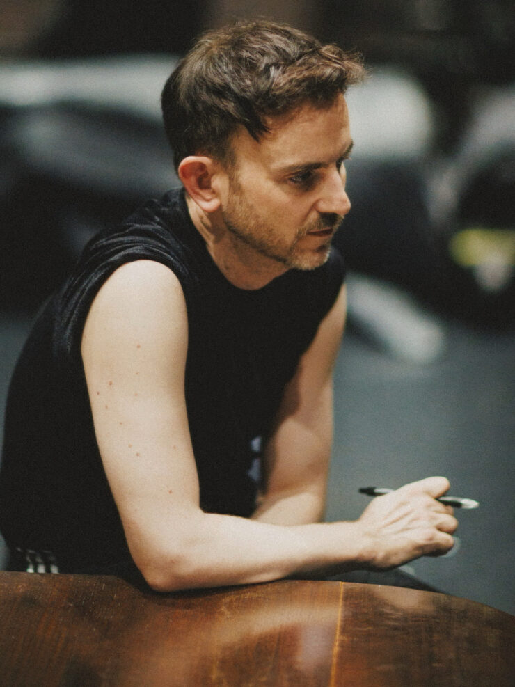 A man leanding on a desk, holding a pencil, watching intently.