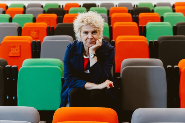 Woman with white curly hair staring out at the camera, with her hand under her chin. She sits amidst colourful theatre seats.
